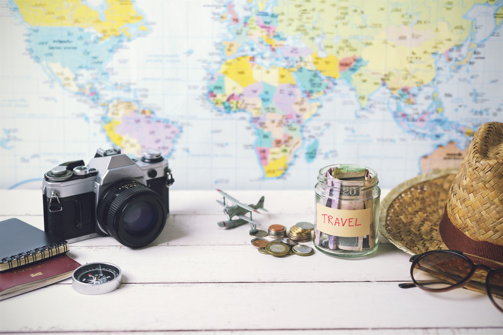World map with a film camera, coins, a travel savings jar, a compass and a straw hat